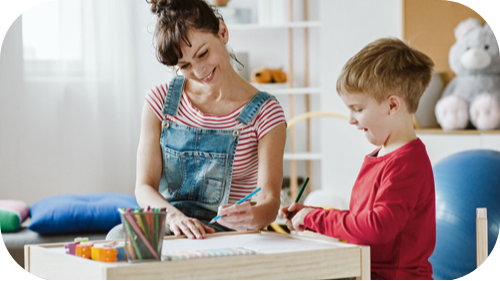 Young boy with mother drawing or writing on a desk with paper.